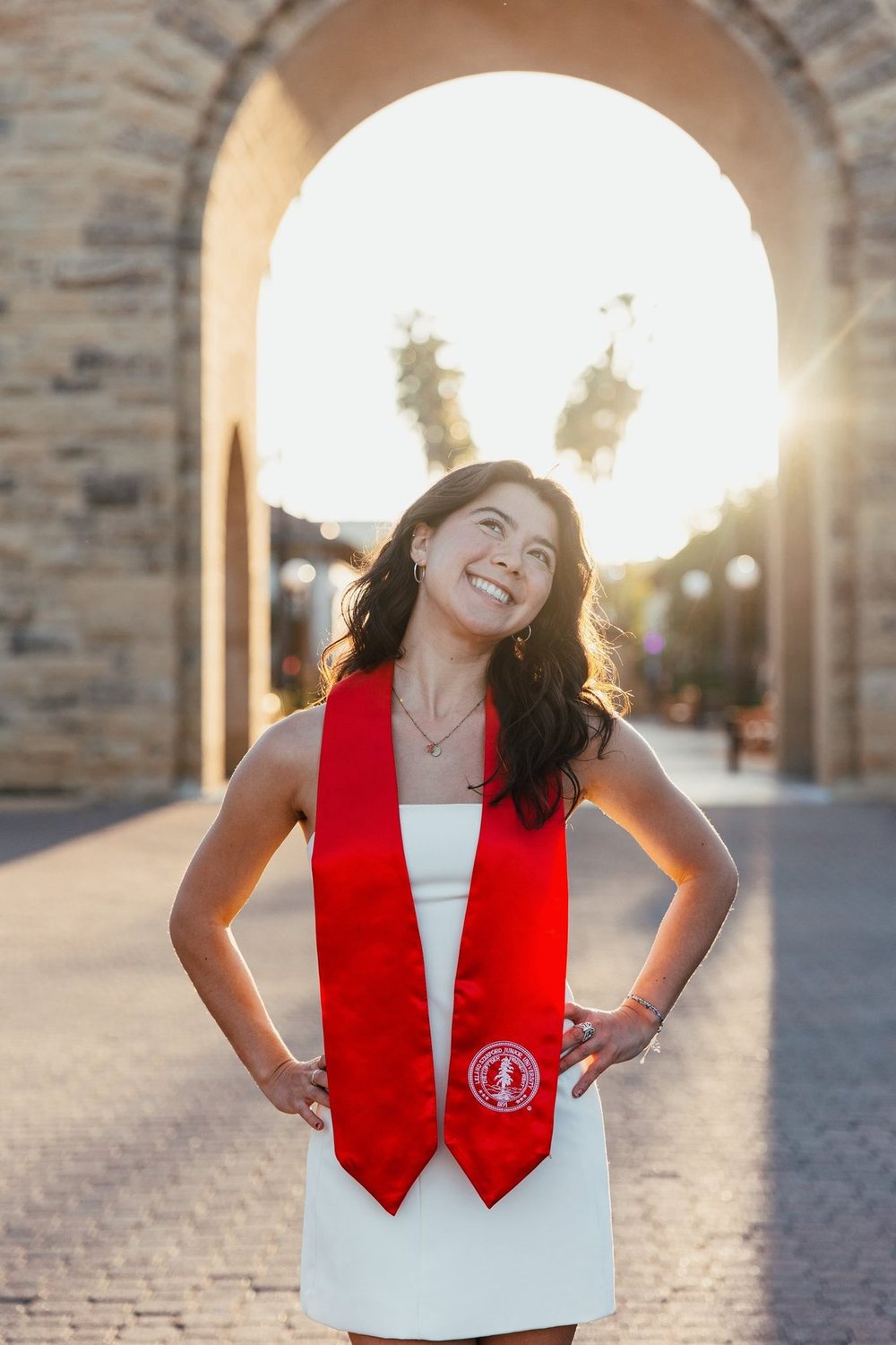 Symphony in a white dress and red Stanford graduation stole, smiling under a sandstone arch